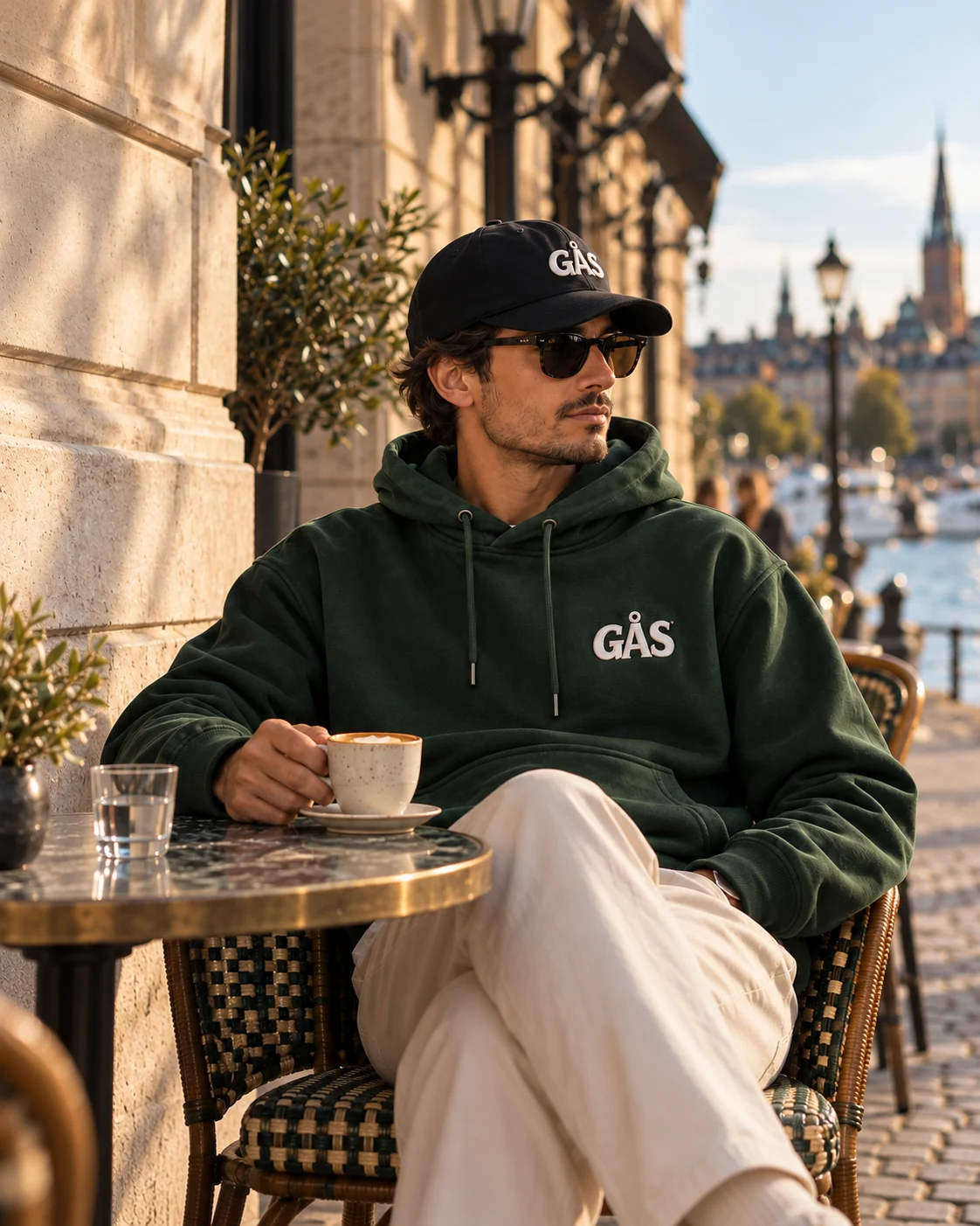 Man i grön GÅS-hoodie och svart keps vid ett café, designmockup